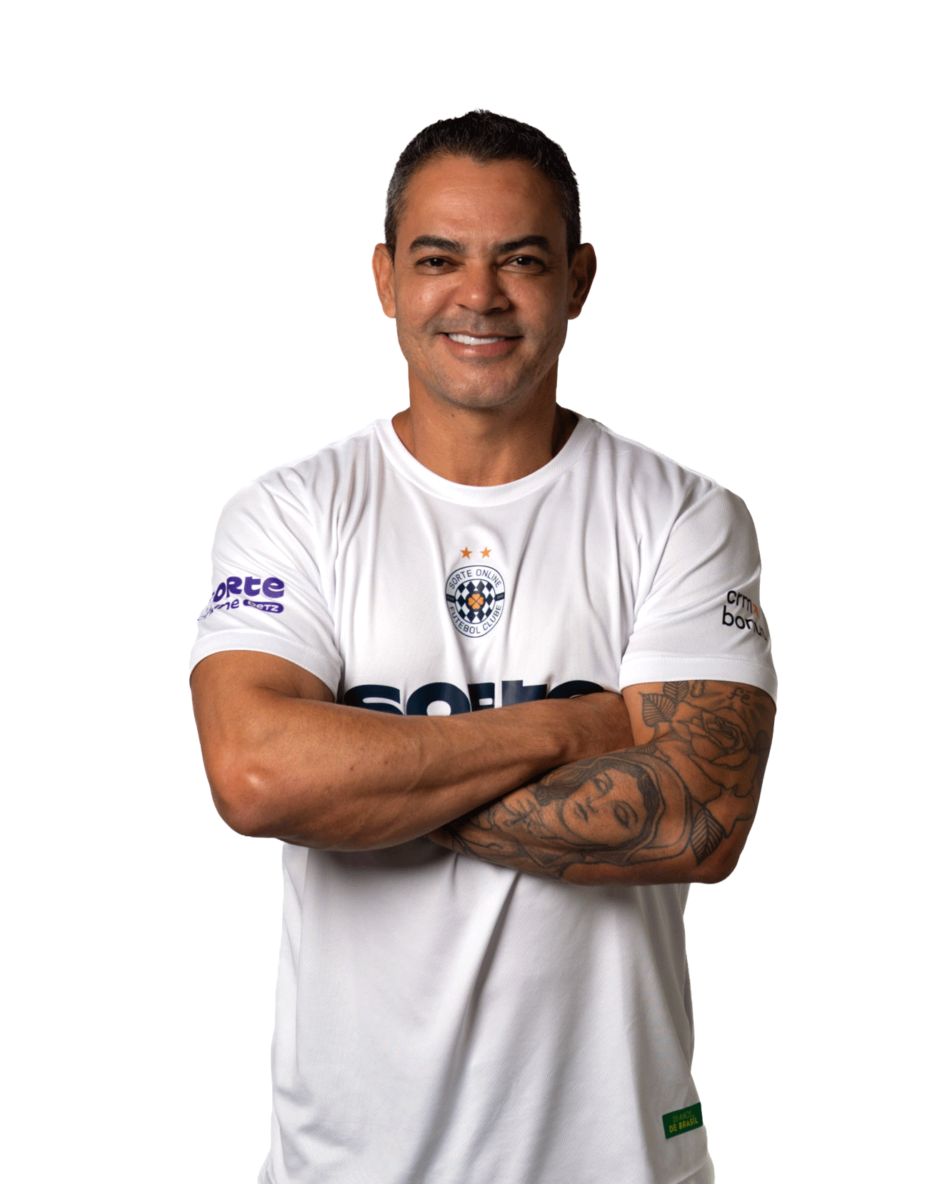 Josué Anunciado de Oliveira, ex-jogador de futebol sorrindo com os braços cruzados, usando camiseta branca do clube Sorte Online Betz com logo e patrocinadores.