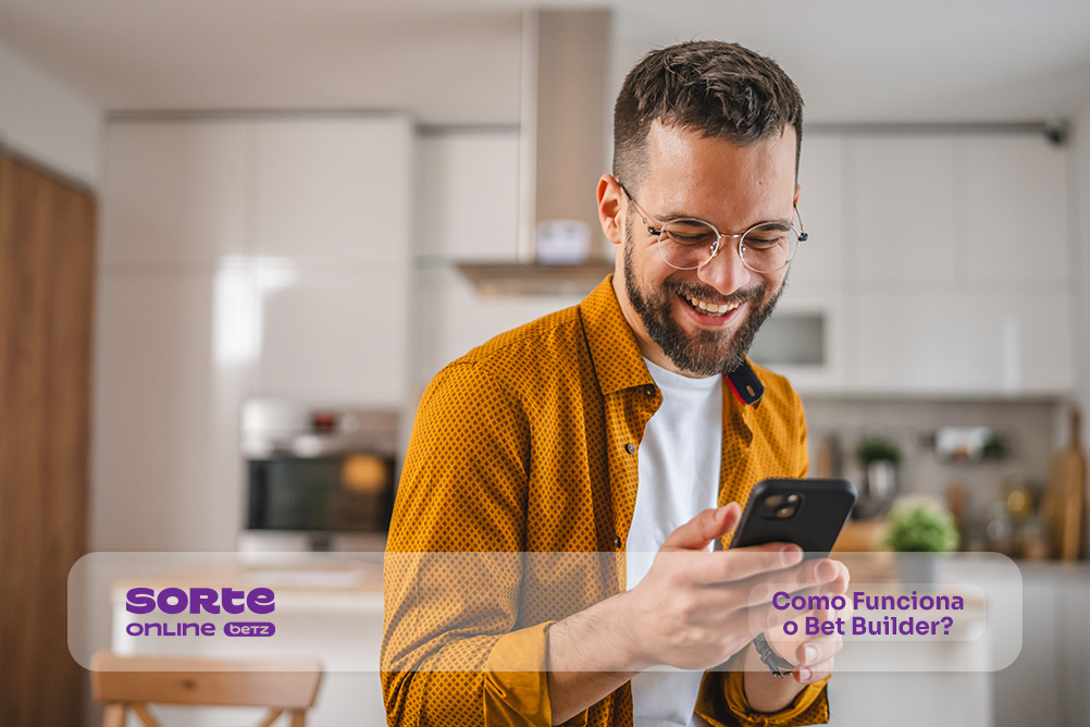 Homem sorrindo e olhando para o celular em sua mão, cozinha embaçada ao fundo. Sorte Online Betz explica como funciona o Bet Builder (Criador de Aposta).
